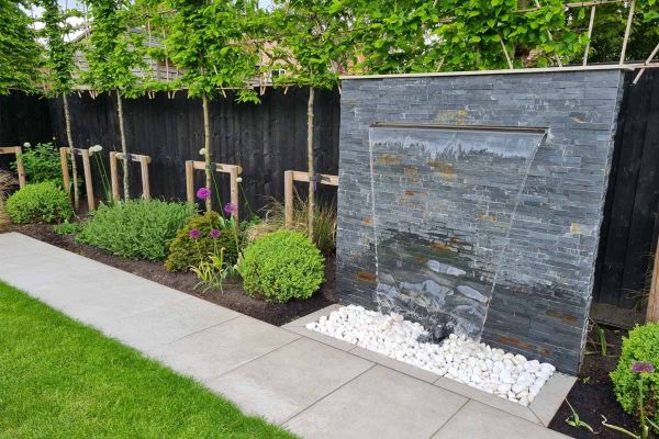 Water pours from black slate cladding waterfall feature into white pebbles with porcelain path running past.***Andrew Jordan Landscape & Garden Design