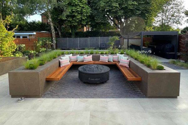 Patio with raised flowerbeds used as walls to contain floating bench and firepit, sitting on lugano clay pavers. Pergola in distance.***Trendy Landscapes