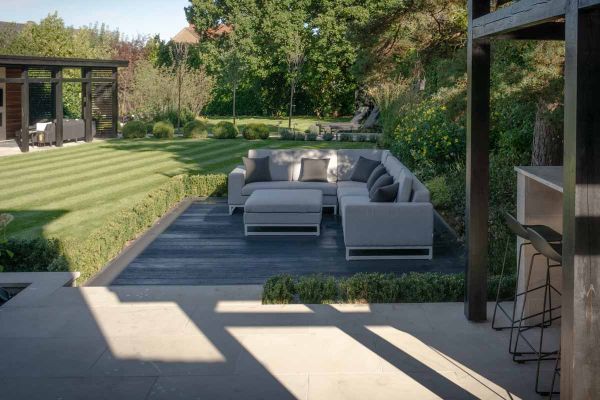 Sun casts shadow over burnt cedar millboard decking area, hosted by large corner outdoor sofa with well manicured lawn in the background.***Robert Hughes Garden Design, www.roberthughesgardendesign.co.uk
