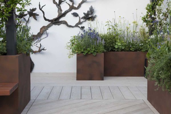 In front of white wall with metal sculpture attached, corten steel planters sit on Limed Oak Millboard decking laid in 3 directions.***Designed by Rhiannon Williams | Built by Garden Club London Ltd, www.gardenclublondon.co.uk