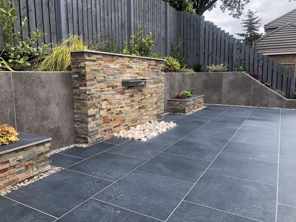 Charcoal porcelain paving patio in front of a stone clad water feature and large retaining wall clad with large format porcelain cladding.***Manor Prestige Landscapes, www.manor-prestige-landscapes-ltd.business.site