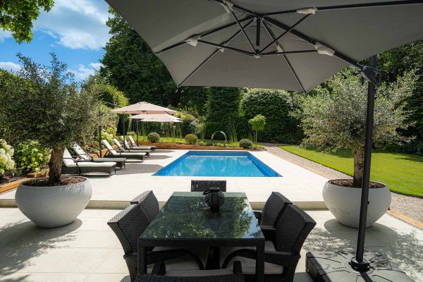 Sun shade covers glass top table with large swimming pool that uses light grey porcelain 20mm bullnose coping stones, and sunloungers in the distance.***J Freeman Builds, www.jfreemanbuilds.co.uk