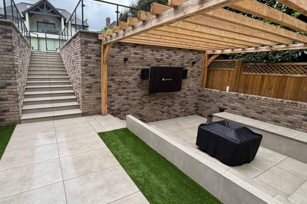 Outdoor entertainment space beneath a timber pergola, finished with Grey York Porcelain Paving and built-in seating.***Bjorn Build 
