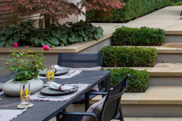 Dining table sits at the bottom of 3 britannia buff yorkstone steps, with pencil round coping stones topping the raised flowerbeds.***Harrington Porter, www.harringtonporter.com
