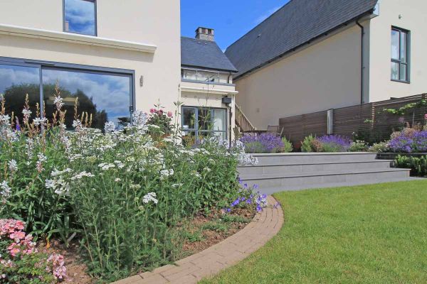 White house with french doors has patio leading down with steel grey porcelain 20mm bullnose steps towards lawn and curved flowerbeds.***Outerspace Creative Landscaping