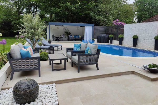 Swimming pool terrace with oval pool, seating area, sun loungers and open sided pergola. Paved with rectangle porcelain paving and bullnose steps.***20mm Bullnose | Chloe Luxon Garden Design |  Executive Gardens