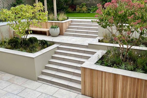 Steps flanked by trees in beds rise in 2 consecutive flights from paving to upper garden level. Jura Grey smooth coping caps walls.***5mm Chamfer Collection | Tom Howard Garden Design
