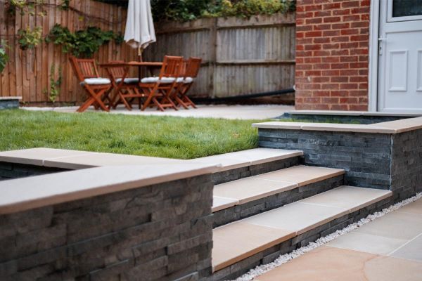 3 steps of Antique Cream sawn paving slabs rise between flanking stone clad walls to lawn next to patio in fenced garden.***Vu Garden Design,  www.vugarden.co.uk