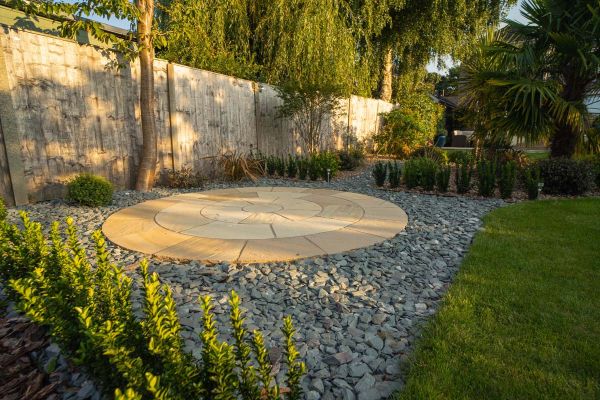 Dappled sunlight falls on Mint sandstone circle set next to tall fence into square of grey gravel with low hedge border.***Austin Landscapes,  www.austinlandscapes.co.uk