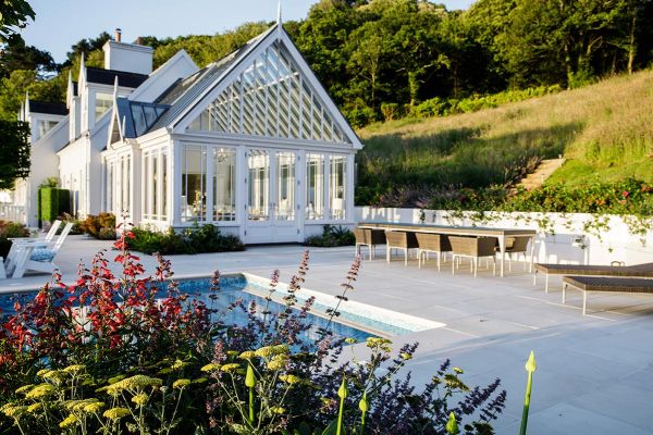 Grass slopes down behind large area of Beige smooth sandstone patio paving between white-painted conservatory and swimming pool.***The Lovely Garden, www.thelovelygarden.co.uk