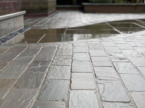 Low view of wet Kandla grey cobble setts with small puddle, showing riven surface. Design by Brockstone Landscape Construction.***Brockstone Landscape Construction Ltd, www.brockstone.co.uk