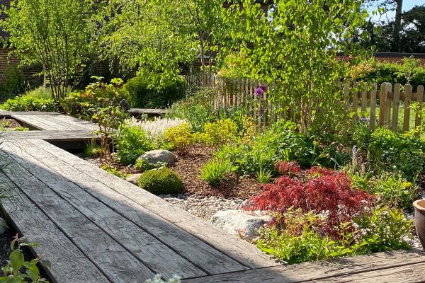 Zigzag boardwalk of Driftwood Millboard decking passes gravel-mulched bed with low shrubs and picket fence. Design by Bushy Business.***Bushy Business, www.bushybusiness.com