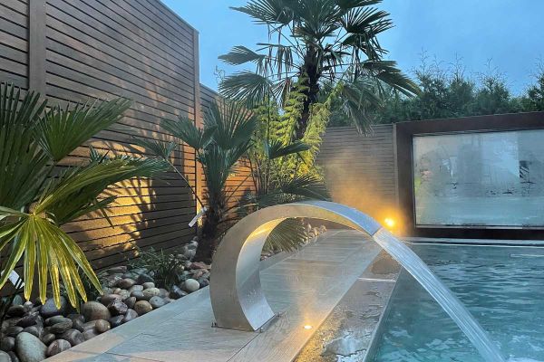 C-shaped water feature shoots flume into swimming pool in front of pebble-mulched bed at base of fence of Warm Teak composite wood.***(image shows wet battens) Evergreen Gardens, www.evergreengardens.biz