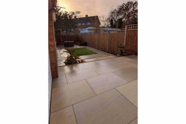 Long narrow fenced plot with wet patio of Raj Green porcelain garden tiles UK and 2 steps down to lawn framed by path. ***Designed & Built by The Shed London Landscapes