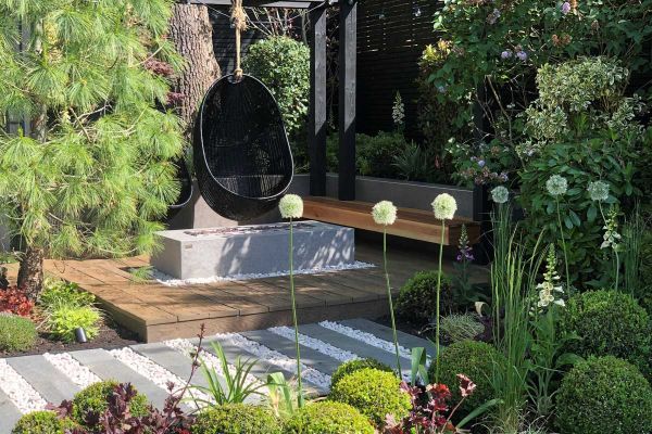 Black egg chairs hang from wooden pergola over vintage millboard decking, with firepit, bench and lush planting surrounding.***Gardens Of The Future, www.gardensofthefuture.co.uk
