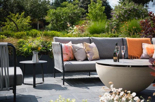 Grey-cushioned garden sofa and table on Brazilian Black stone slabs with lush planted beds in large garden behind.***Designed by Greencube, www.greencubelandscapes.co.uk | Built By Langdale Landscapes,  www.langadalelandscapes.co.uk
