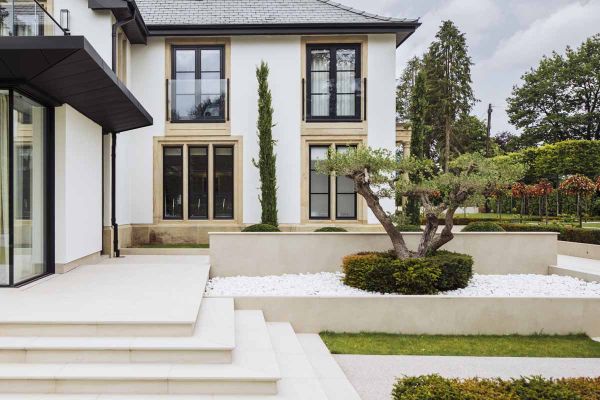 French-looking house with wrap-around patio with steps descending next to raised bed faced in Off White Premium DesignClad.***Designed by Luxe Exteriors, www.luxeexteriors.co.uk | Built by Green Acorn Garden Design, www.greenacornlandscapedesign.co.uk
