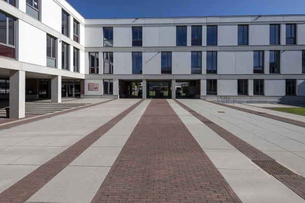 Stripes of Novara clay brick paving alternate with cream-coloured outdoor tiles in large courtyard of 3-storey white building.***