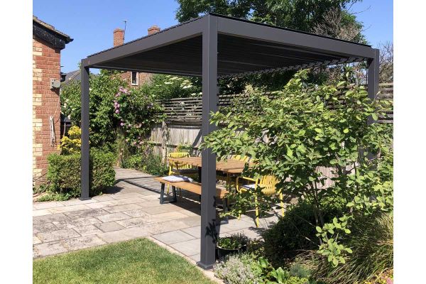 Proteus grey aluminium pergola on sunny patio shelters table with bench and chairs. Large bush in foreground. Design by VaRa.***VaRa Garden Design, www.varagardendesign.co.uk