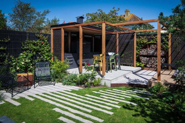 Corner of fenced garden. Light Grey porcelain plank paving set into curved lawn, next to raised patio covered by wooden pergola.***Design by Rachel Goozee Garden & Planting Design, www.rachelgoozee.com | Built by Timeless Landscapes, www.timelesslandscape