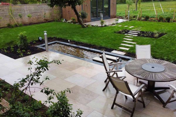 Round wooden table and chairs on top of Raj Green Porcelain Paving overlooks a small pond and large lawn area with outhouse.***LG Landscapes Ltd, www.lglandscapes-trowbridge.co.uk
