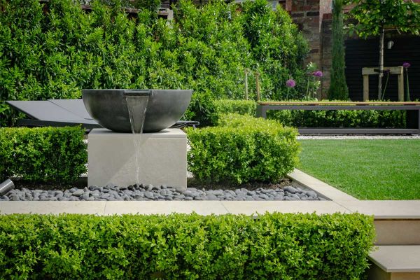 Large bowl spills water onto pebbles in area outlined in Slab Khaki Porcelain Pool Coping with chamfer. Design by Robert Hughes.***Image also displays Paving & Steps | Designed by RobertHughesGardenDesign.co.uk | Built by Brockstone.co.uk