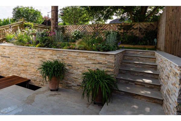Silver grey porcelain slabs for steps rise between stone-clad retaining walls from terrace to decked area under mature trees.***Hythe Garden Landscapes Ltd, www.hythegardenlandscapes.co.uk