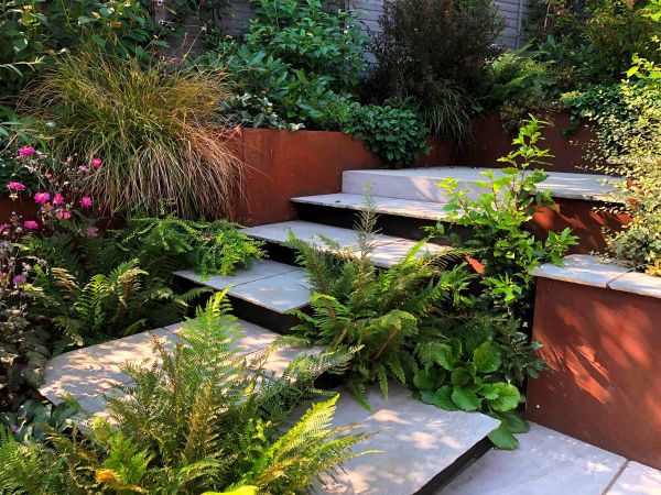 Steps of tumbled Kandla grey garden paving slabs turn right angle and rise to paved area surrounded by lush planting.***VaRa Garden Design, www.varagardendesign.co.uk
