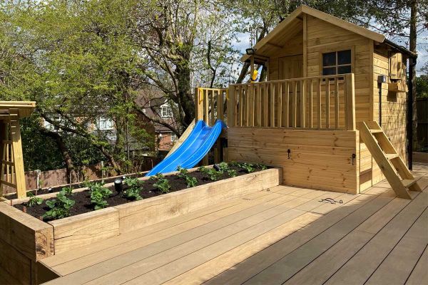 Garden decking of Warm Teak Brushed Designboard with large chalet-style playhouse and raised bed edged with wooden sleepers.***Gardens of the Future, www.facebook.com/gardensofthefutureuk