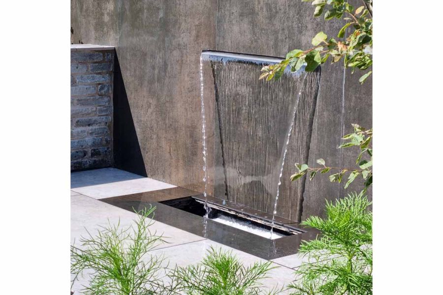 Sun shines on water feature of wall faced with Steel Dark Designclad porcelain cladding with spout gushing into small pond at base.