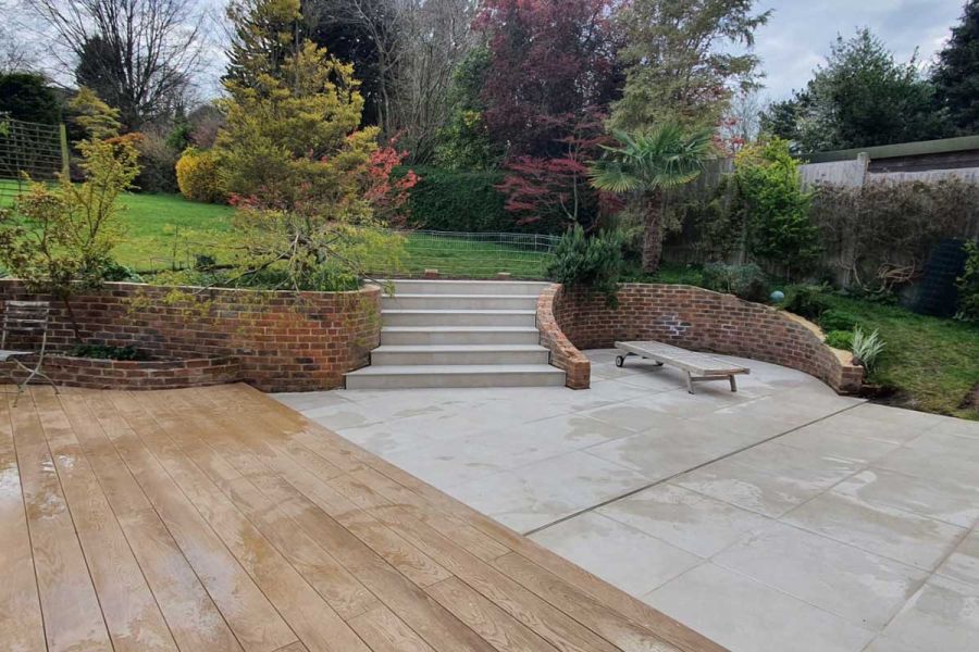 Decking area next to Area 800x800 Porcelain Paving, integrated steps leading up to a lawn, and large paving slabs adjacent to a red brick wall.