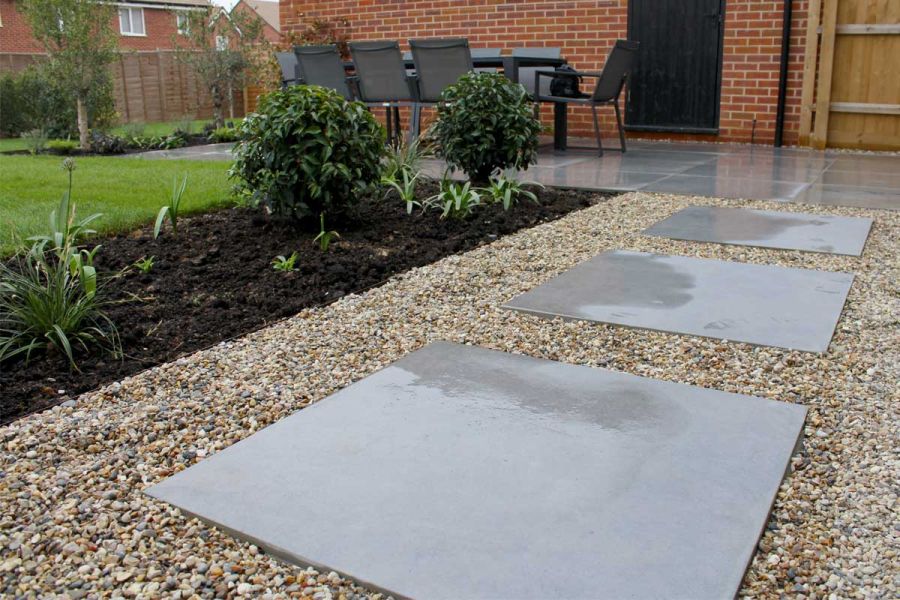 Anthracite 800x800 porcelain paving laid in pea gravel next to bed with small plants. Matching patio with dining set in background.