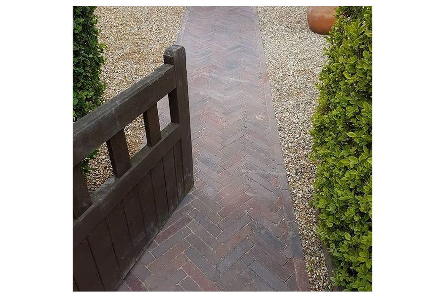 Wooden gate set into hedge opens over straight path of Abbey Dark Multi clay pavers laid herringbone pattern between gravel areas. 