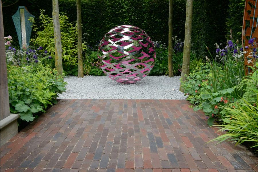 Pink and silver perforated sphere sits on white gravel, approached by wide path of Abbey Dark Multi clay bricks between beds.