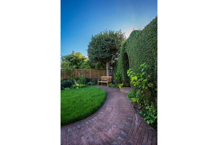 Curved path of Abbey Dark Multi Brick Pavers runs between clipped hedge and lush lawn, past a wooden bench in fenced garden.