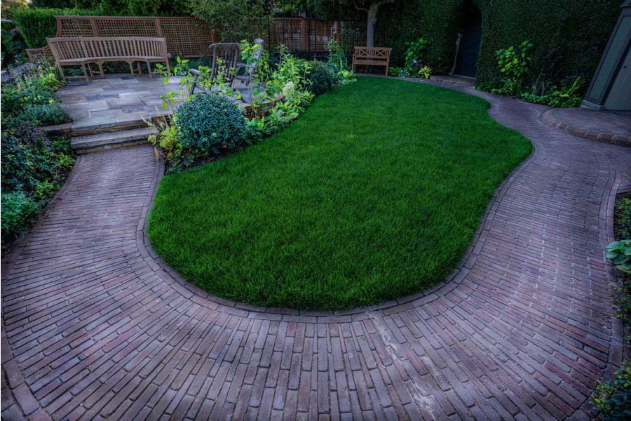 Path of Abbey Dark Multi Brick Pavers curves between hedge and lawn, past a wooden bench in fenced garden. Design by Jo Thompson.