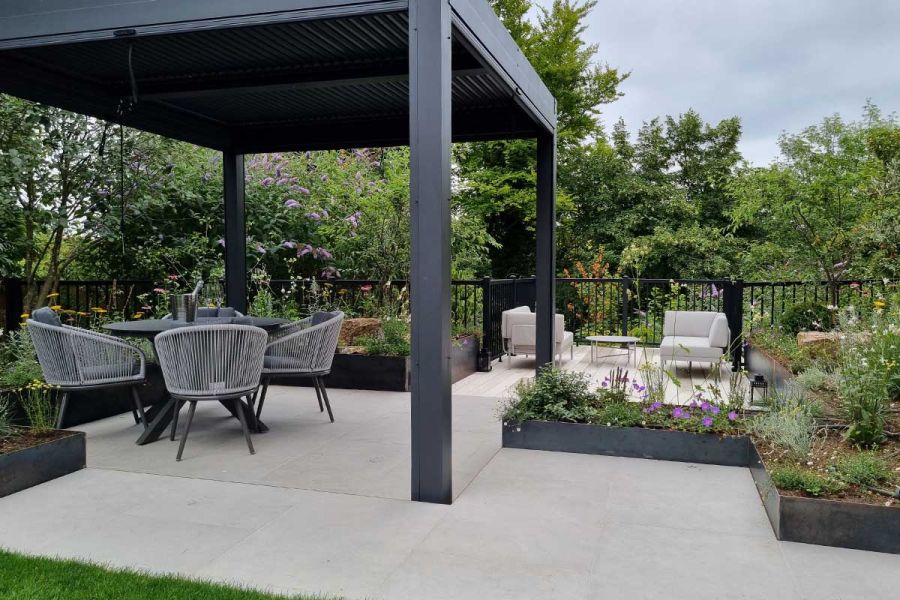 Large metal pergola covers dining area, next to raised beds and railed seating area. All paved in Florence White porcelain slabs.