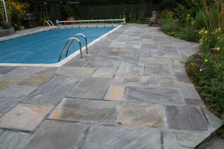 Large rectangular swimming pool with wide Tumbled Black sandstone paving surround, edged with planted border and fence.
