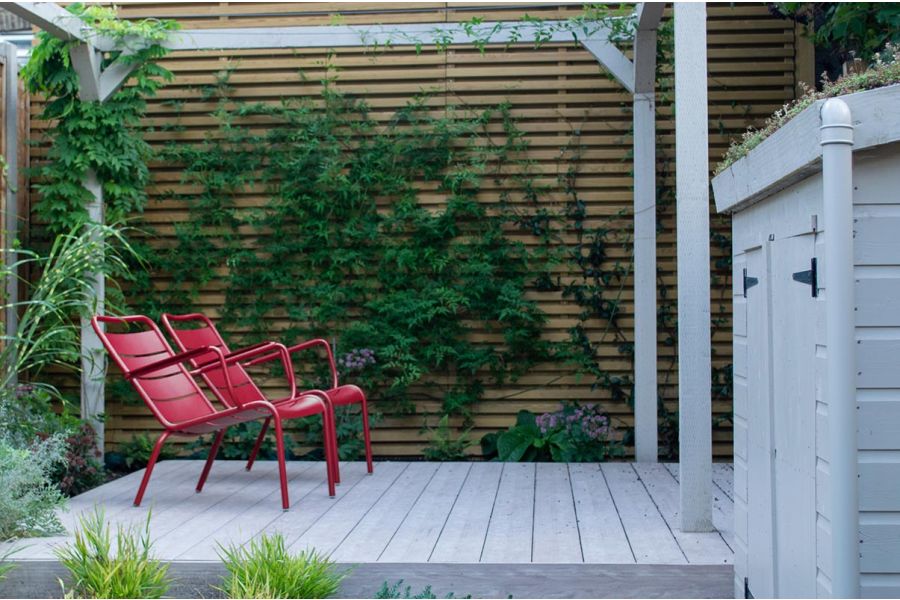 2 red metal chairs on Cinnamon composite decking under matching pergola, next to storage shed. Design by Alexa Ryan-Mills.