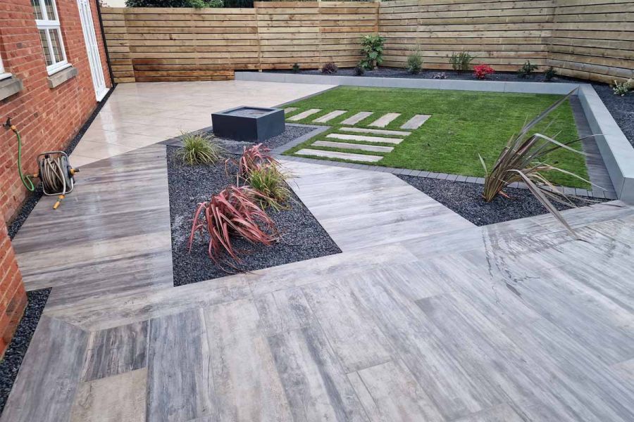 Fenced back garden with geometric layout of Cinder porcelain paving, lawn and grey gravel-mulched beds. Design by All Seasons.