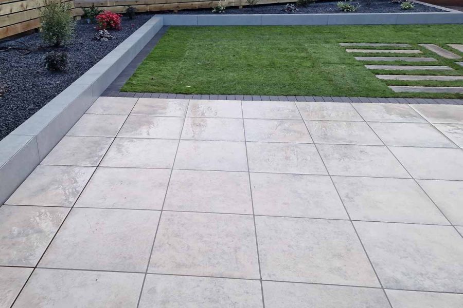 Wide mulched bed with kerb edge runs alongside oblong lawn with stepping stone path and large patio of Jura Grey porcelain paving.