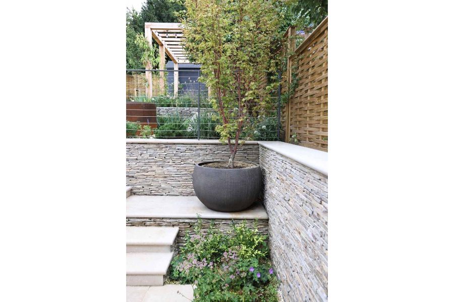 View from the corner of a garden, a large plant pot sits on Jura Beige bullnose steps, clad with cobblestones and surrounded by plants.
