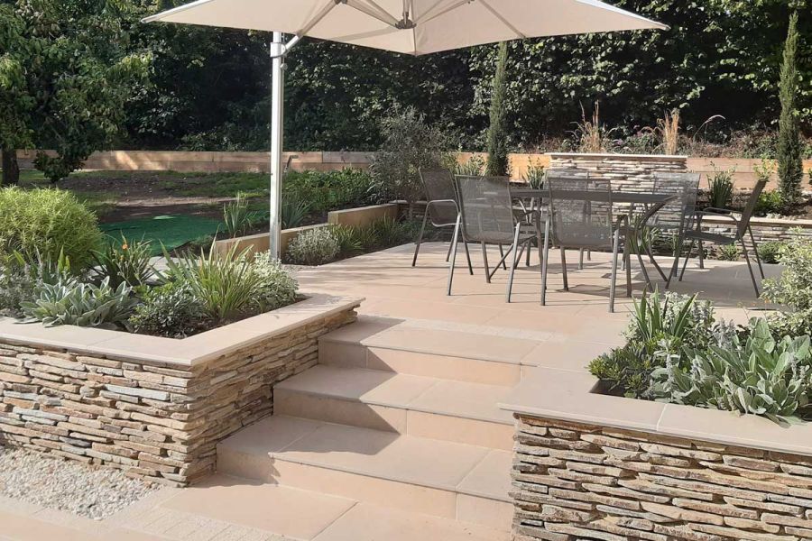 Florence beige 5mm chamfer porcelain garden steps rise to patio with stone-clad walls and planted beds. Design by Anna Helps.