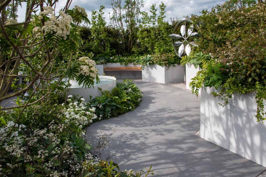 Wide path of Anthracite Porcelain Large Patio Slabs curves round central raised pond bordered by green and white planted bed.