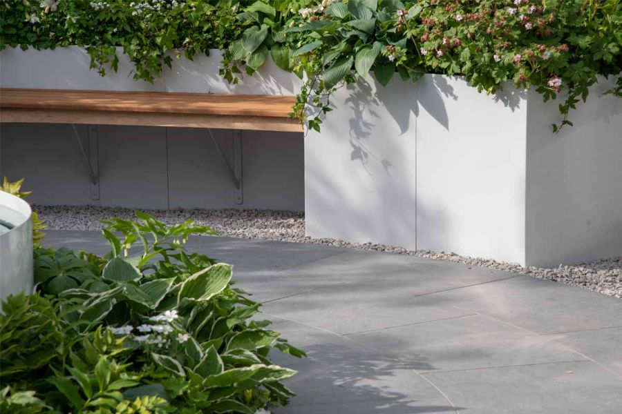 At RHS Malvern Flower Show, path of Anthracite Porcelain paving at base of curved white-walled raised bed with inset wooden bench.