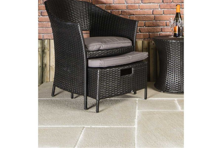Rattan armchair with footstool sits on Antique Cream sandstone smooth paving slabs with pale mortar joints, in front of brick wall.