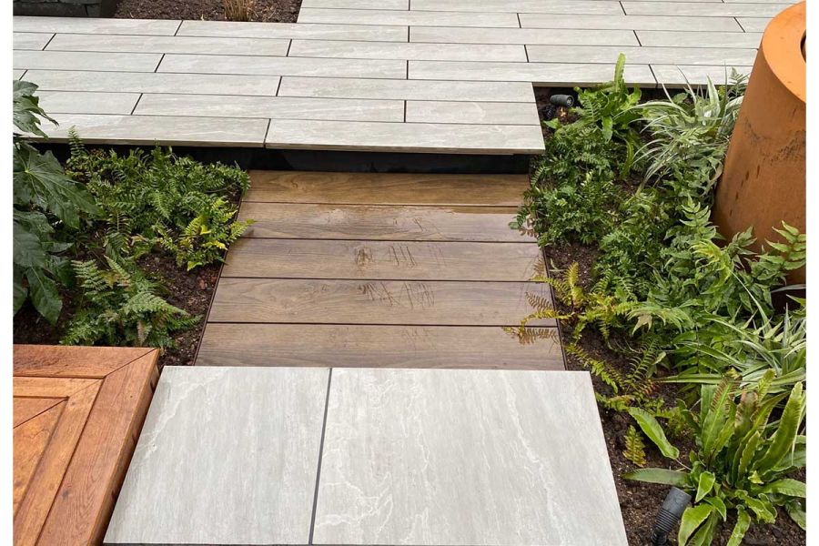 5 short Antique Oak Millboard decking planks, laid flush with planted beds on either side, divide 2 areas of porcelain paving.