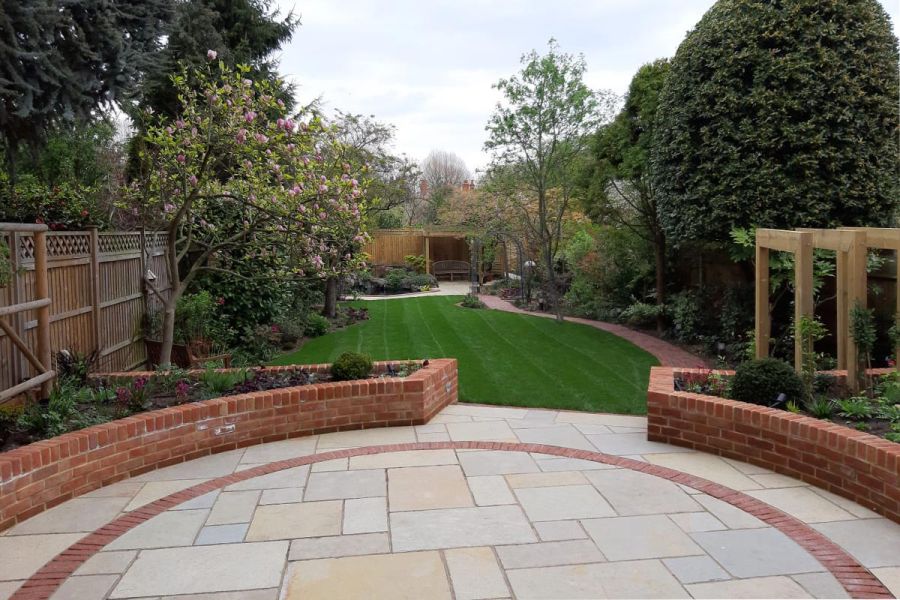 2 curve-edged beds form corners of Antique Yellow limestone patio with curved brick paver detailing in fenced garden with lawn.