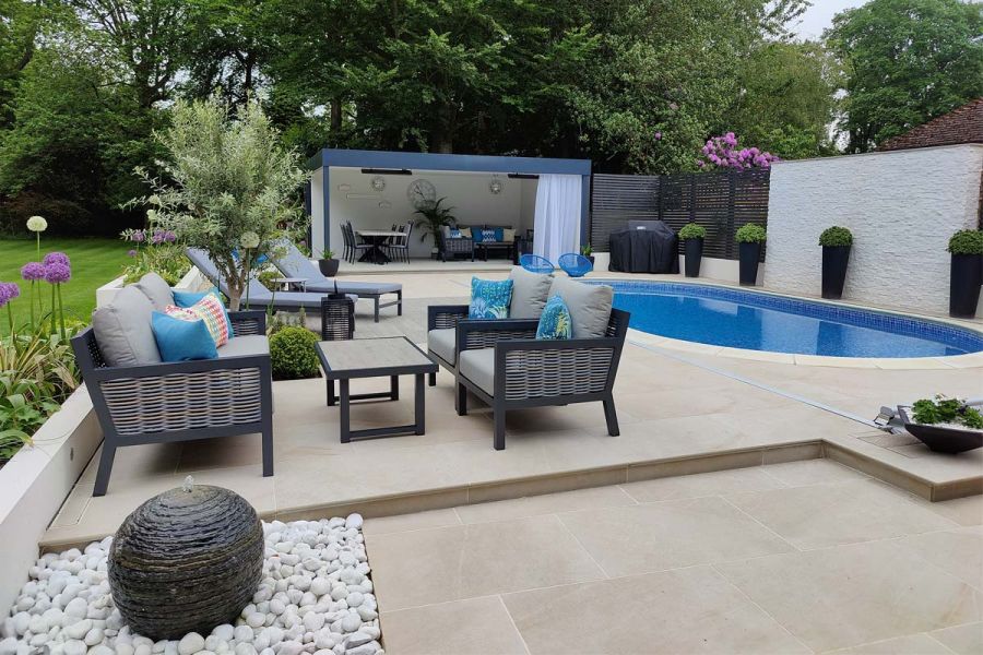 Swimming pool terrace with large oval pool, seating area, sun lounger and a large open sided pergola. Paved with rectangle porcelain paving.
