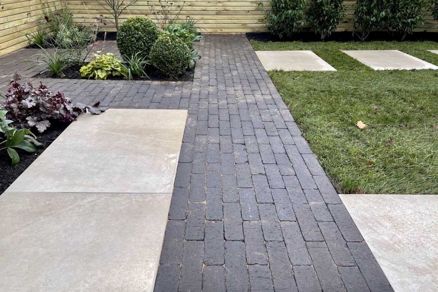 Paved garden area made up of a mix of clay pavers and Porcelain paving slabs interspersed with planted pockets and leading towards a lawn.